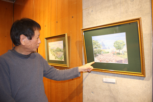 ＜写真：27日、水彩画「仁摩サンドミュージアム」の前で映る松本さん＝大田市仁摩町＞