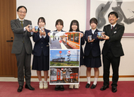 〈写真：新しく開発したラベルのポスターを紹介する平田高校の生徒たちと、新ラベルの商品を手にする持田祐輔社長（左）と一畑電車営業主任・電車運転士の宅野康平さん（右）＝25日、出雲市今市町の出雲市役所〉
