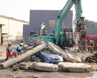 〈写真：大型重機によるがれき撤去訓練を行う、山﨑組社員と大田消防署員＝大田市鳥井町の山﨑組〉 