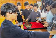  〈写真：藤井重綺さん（左）のチャンピオンベルトに目を輝かせる児童たち＝14日、出雲市立塩冶小学校〉